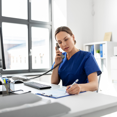 Female nurse on the phone writing down on a clip board while sitting at a computer
